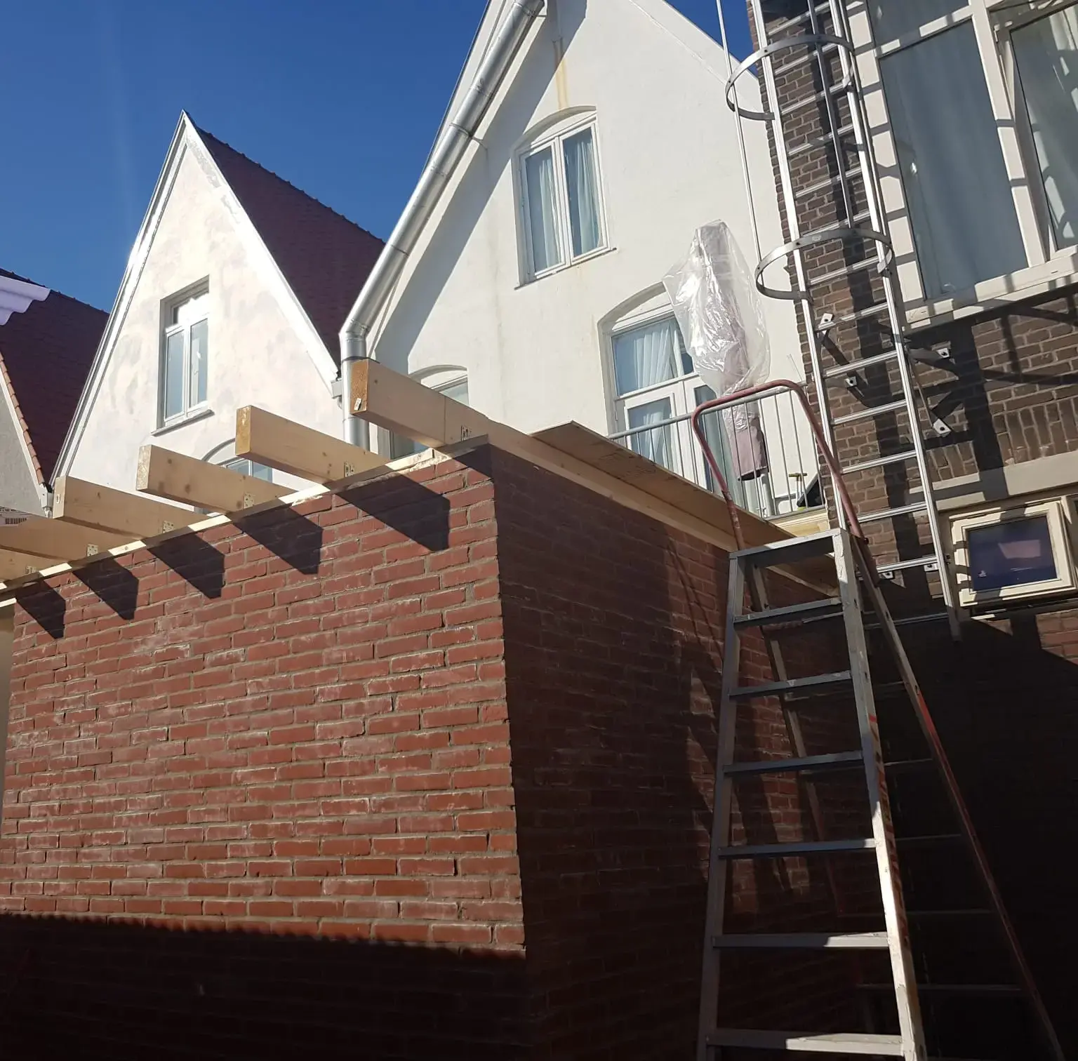 Gemetselde aanbouw achter een woning met puntgevels; de houten dakbalken liggen erop en er staat een ladder tegenaan.